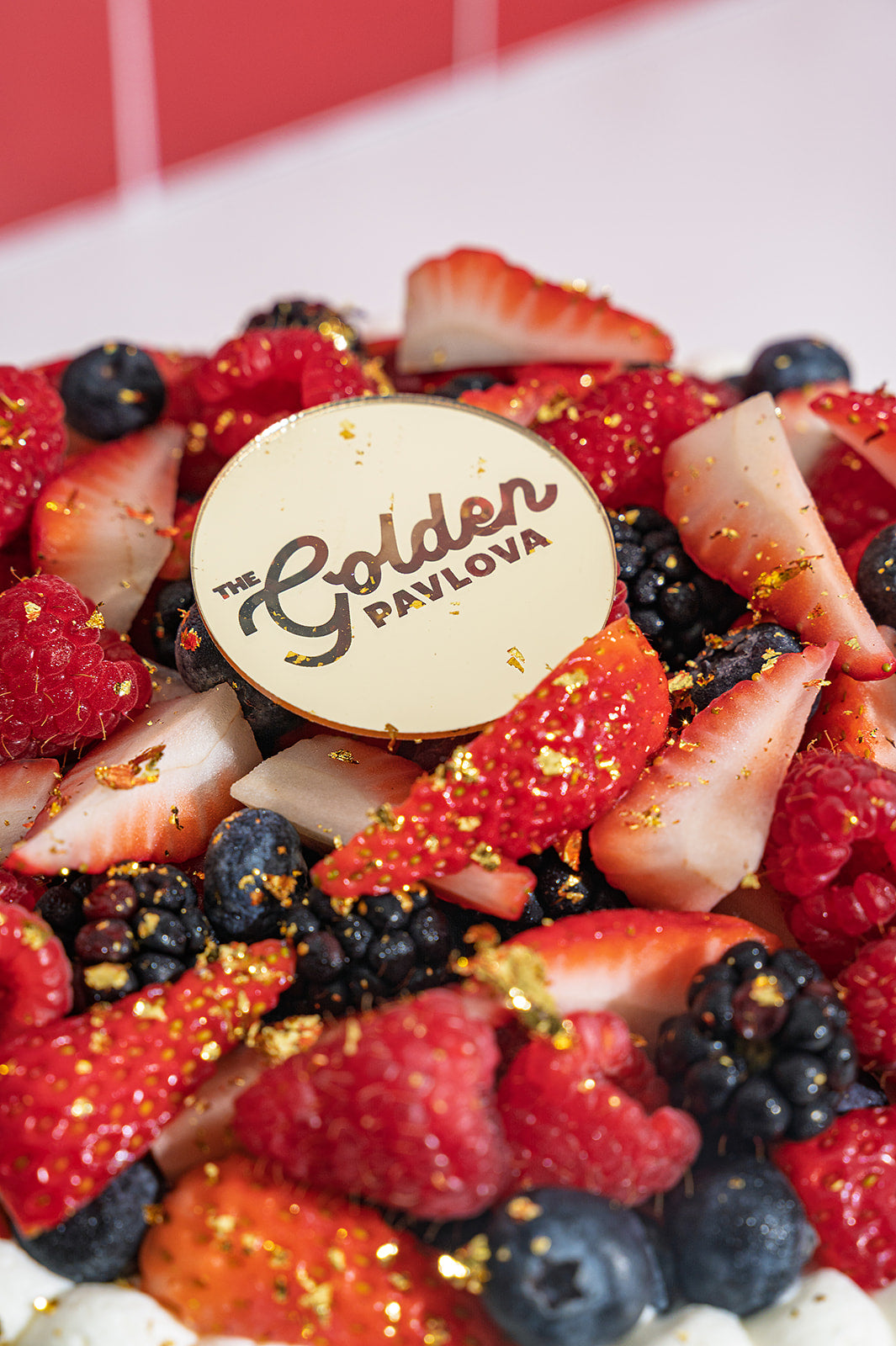 Close-up of mixed berries pavlova with fresh strawberries, blueberries and raspberries, finished with golden flakes and The Golden Pavlova logo.